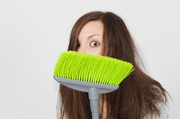 Cerca de la diversión joven loca ama de casa asustada salvaje loca mareada con el pelo despeinado aislado sobre fondo blanco. Mujer loca ama de llaves que se esconde detrás de la escoba verde. Copie el espacio para publicidad.