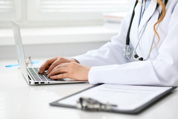 Cerca de una doctora desconocida sentada en la mesa cerca de la ventana del hospital y escribiendo en una computadora portátil