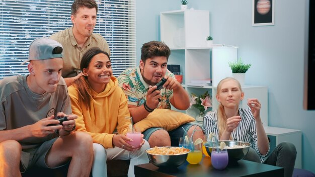 Foto cerca de dos chicos jugando videoconsola, uno de ellos gana. sus amigos los animan, se sientan en el sofá en una bonita casa, comen bocadillos y se divierten.