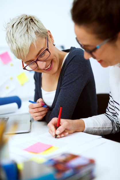 Cerca de dos sonrientes mujeres de mediana edad de negocios innovadores y elegantes que trabajan juntos en la resolución de problemas mientras están sentados en la oficina uno al lado del otro.