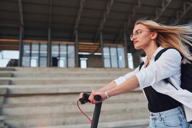 Cerca del edificio de negocios Hermosa rubia con ropa informal montando una moto eléctrica al aire libre durante el día soleado