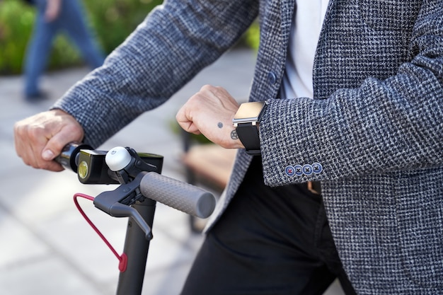 Cerca del elegante empresario de mediana edad comprobando el tiempo mientras está de pie con scooter eléctrico