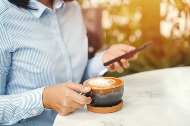 Cerca de la empresaria utilizando teléfono inteligente y bebiendo café con leche caliente en la mañana en el café.