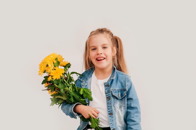 De cerca, enfoque selectivo. Niña rubia en un sostiene crisantemos y gerberas en sus manos sobre un fondo blanco, una niña sonríe y sostiene flores de primavera en sus manos