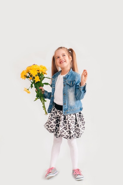 De cerca, enfoque selectivo. Niña rubia en un sostiene crisantemos y gerberas en sus manos sobre un fondo blanco, una niña sonríe y sostiene flores de primavera en sus manos