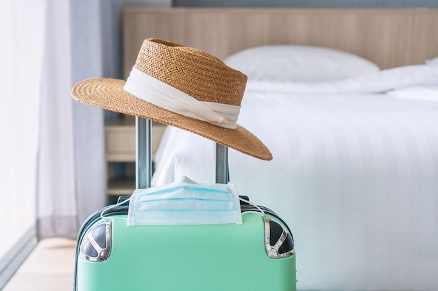 Cerca de equipaje verde, sombrero de paja y mascarilla quirúrgica. Viajando en un nuevo concepto de estilo de vida normal.