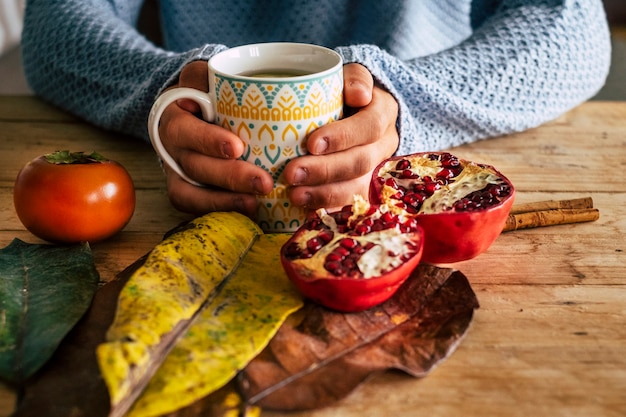 Cerca de la escena interior de la casa de otoño con un par de manos sostenga una taza de té en una mesa de madera de color amarillo y naranja con pomelo y hojas