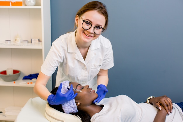 Cerca de esteticista cosmetóloga inyectando botox en mejillas femeninas. Retrato de atractiva chica africana y cosmetóloga en guantes. Esteticista tocando la frente de la dama y haciendo inyección