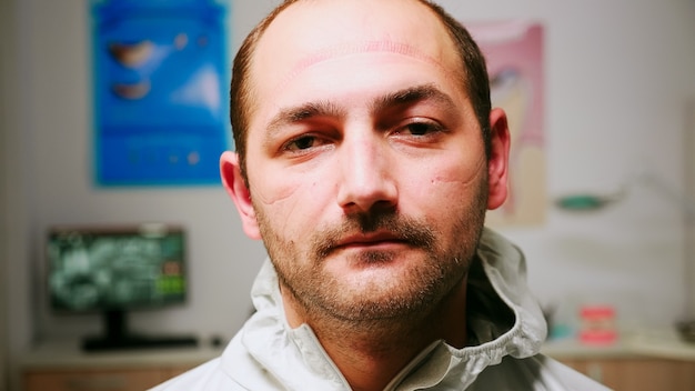 Cerca del estomatólogo pediátrico cansado hombre despegando el protector facial mirando a la cámara agotado sentado en el nuevo consultorio dental normal. Equipo médico con traje y guantes de ppe