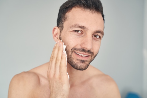 Cerca de feliz hombre barbudo con cuerpo musculoso en el momento de aplicar la crema de afeitar en el rostro