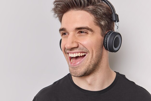 Cerca de feliz hombre positivo sonríe alegremente disfruta de su tiempo libre escucha música en auriculares inalámbricos vestidos casualmente aislado sobre fondo blanco escucha canciones populares utiliza equipos electrónicos