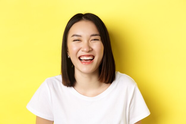 Cerca de feliz joven divirtiéndose, sonriendo y riendo sin preocupaciones, de pie en camiseta blanca sobre amarillo.