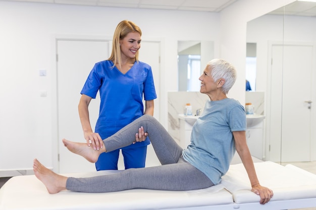 cerca de la fisioterapeuta está ayudando a una mujer mayor estirando su tendón de la cadera y haciendo rehabilitación del muslo o la pierna en la sala de ejercicios ella está acostada en una cama de masaje