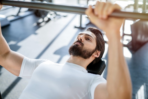 Foto cerca de la formación de hombre fuerte enfocado en gimnasio sosteniendo barra pesada en gimnasio brillante.