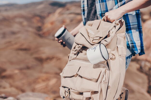 Foto cerca de la foto. las manos de los hombres sostienen un termo y una mochila de senderismo con una taza blanca colgada con una huella. fondo borroso de colinas rocosas. el concepto de turismo.