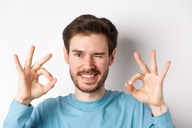 Cerca de guapo hombre barbudo guiñando un ojo y sonriendo, mostrando signos de bien, asegurar que todo esté bien, de pie sobre fondo blanco.