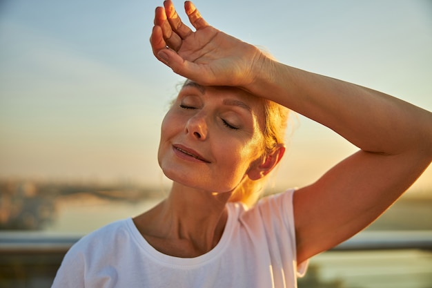 Cerca de la hermosa dama de mediana edad cerrando los ojos y sonriendo mientras descansa su mano en la frente