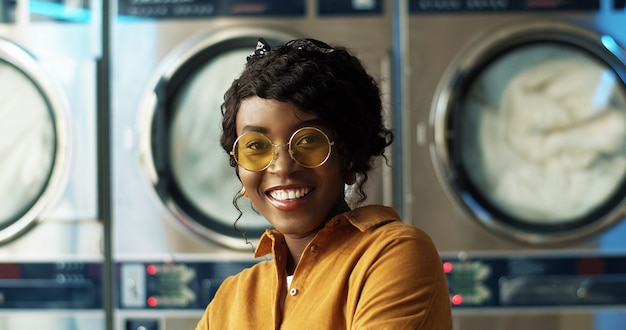 Cerca de la hermosa joven afroamericana en gafas de sol amarillas sonriendo alegremente a la cámara en la habitación de servicio de lavandería. Retrato de niña muy feliz riendo con lavadoras.