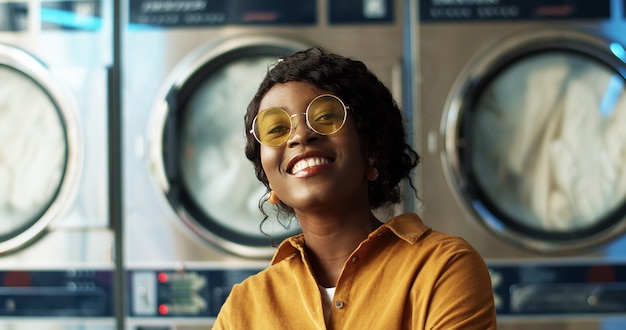 Cerca de la hermosa joven afroamericana en gafas de sol amarillas sonriendo alegremente a la cámara en la habitación de servicio de lavandería. Retrato de niña muy feliz riendo con lavadoras.