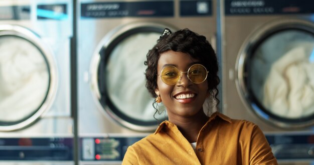 Cerca de la hermosa joven afroamericana en gafas de sol amarillas sonriendo alegremente a la cámara en la habitación de servicio de lavandería. Retrato de niña muy feliz riendo con lavadoras.