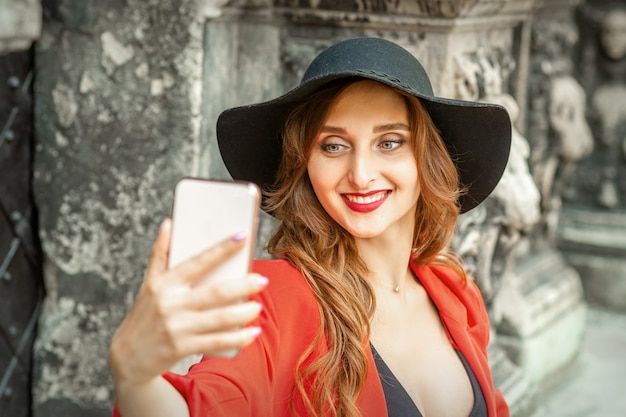 Cerca de la hermosa joven mujer caucásica tomando selfie en smartphone de pie y sonriendo contra el antiguo edificio al aire libre