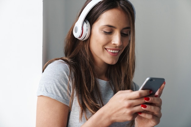 Cerca de una hermosa joven sonriente de pie apoyado en una pared gris sobre una pared gris, escuchando música con auriculares inalámbricos mientras sostiene el teléfono móvil
