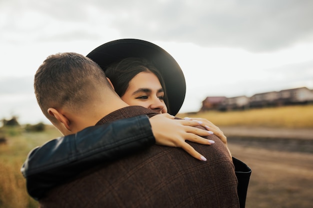 Cerca de una hermosa joven sonriente con sombrero negro abrazando a su novio y abrazándolo con fuerza.