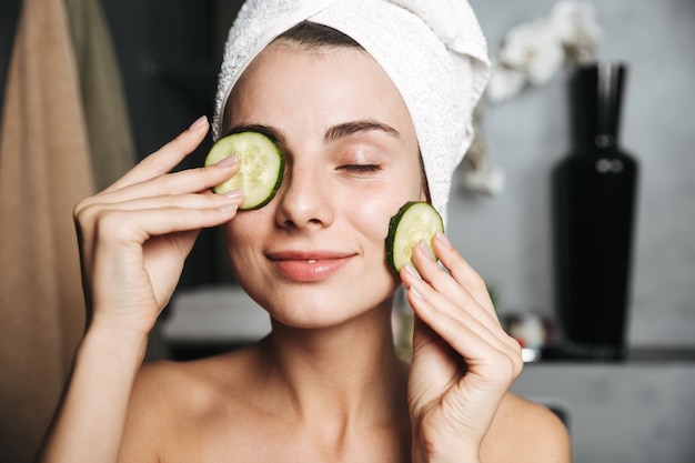 Cerca de una hermosa joven con una toalla envuelta alrededor de su cabeza sosteniendo pepino en rodajas en su cara en el baño