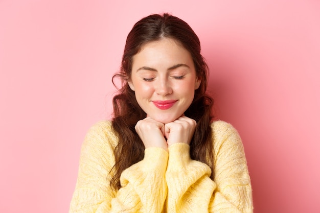 Cerca de la hermosa mujer morena soñando despierto, cerrar los ojos y sonriendo, pensando en el momento romántico, feliz de pie contra la pared rosa en pose tierna.