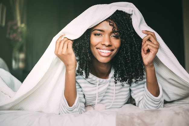 Cerca de una hermosa mujer negra con cabello rizado sonriendo y cubriendo con sábanas en la cama mirando a la cámara