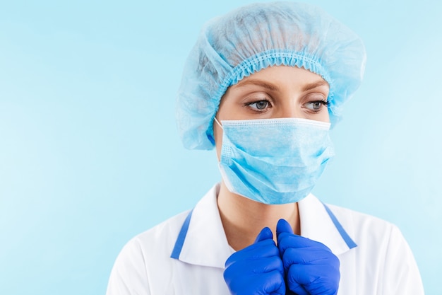 Cerca de una hermosa mujer rubia médico con uniforme y máscara que se encuentran aisladas sobre la pared azul, mirando a otro lado