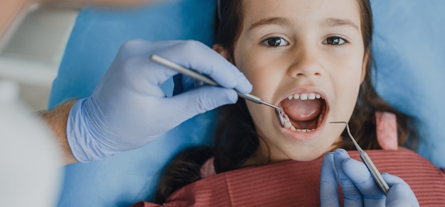 Cerca de una hermosa niña sentada en un asiento de estomatología con un examen dental por un estomatólogo pediátrico.