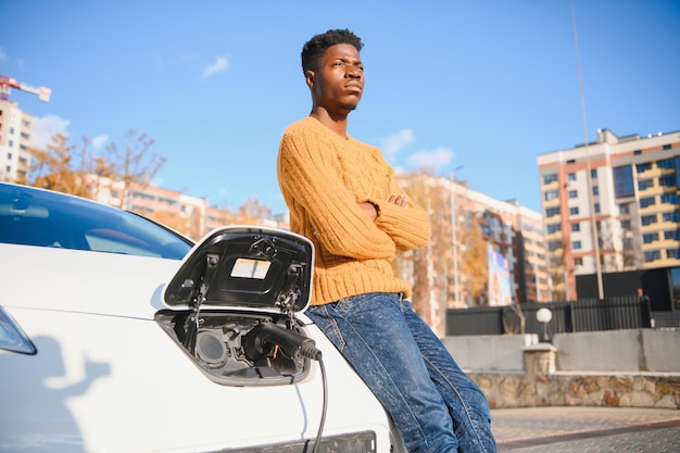 Cerca del hombre afroamericano que conecta el cable de carga al coche eléctrico. Varón joven parado cerca de su auto moderno con maleta de cuero en mano.