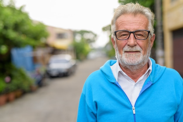 Cerca de hombre barbudo senior guapo feliz sonriendo mientras usa anteojos y chaqueta azul al aire libre