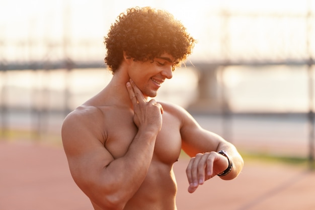 Cerca del hombre deportivo sonriente sin camisa que controla el latido del corazón después de correr