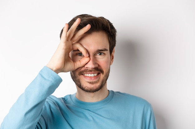 Cerca de hombre feliz mirando a través de signo bien y sonriendo, aprobar y como algo bueno, de pie sobre fondo blanco.