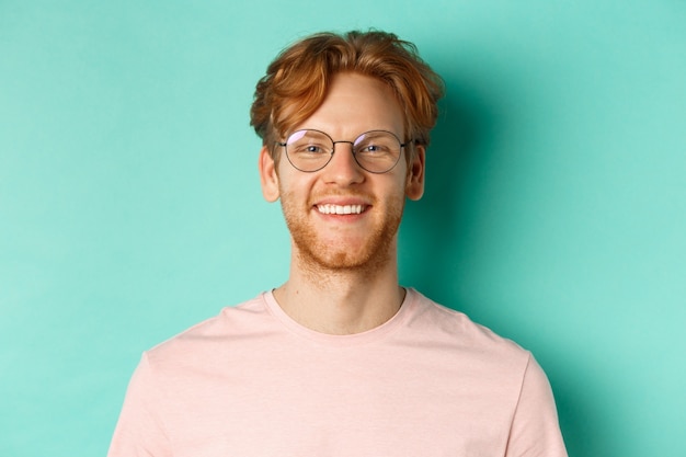 Cerca de hombre guapo pelirrojo con gafas mirando a cámara, sonriendo con dientes blancos, de pie contra el fondo de menta.