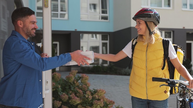 Foto cerca de un hombre guapo que abre la puerta a una repartidora de pizza con una mochila térmica amarilla en una bicicleta y recibe cajas de cartón de pizza concepto de entrega de alimentos