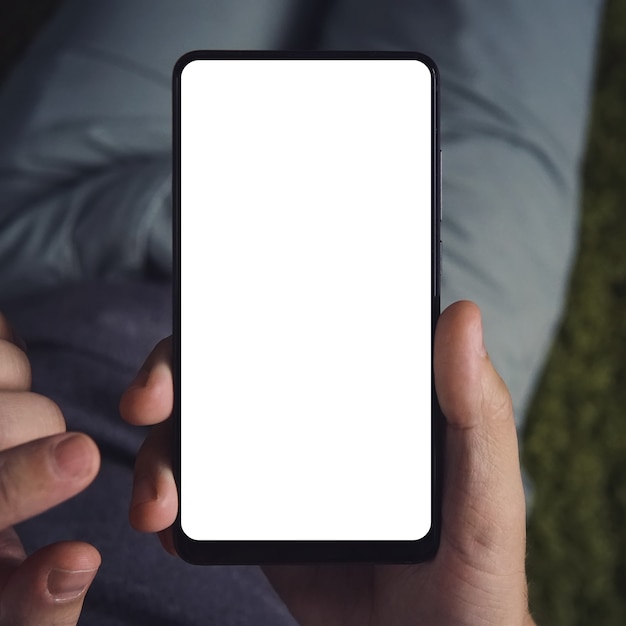 Cerca de un hombre joven sentado durante mucho tiempo sosteniendo el teléfono inteligente con la pantalla en blanco. La persona está en línea desde un teléfono inteligente. Cerca de un hombre usa el teléfono celular. Pantalla en blanco para montaje de visualización de gráficos.