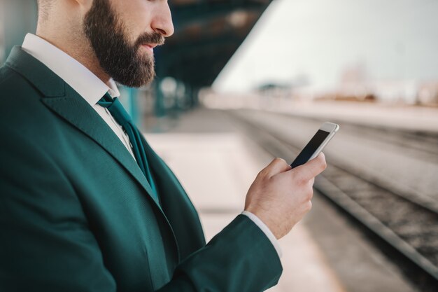 Cerca del hombre de negocios caucásico barbudo serio en traje de color turquesa con teléfono inteligente para leer o escribir mensajes mientras espera el tren. Solo sigue apareciendo cuando la mayoría de las personas renuncien.