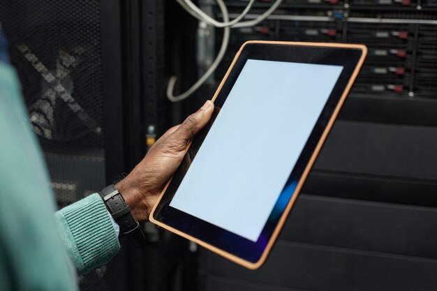 Cerca del ingeniero de datos afroamericano que sostiene la tableta digital con pantalla en blanco mientras trabaja con la supercomputadora en la sala de servidores iluminada por la luz azul, espacio de copia