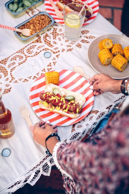 Cerca de irreconocible joven sosteniendo placa de rayas con hot dog americano y maíz en la fiesta de verano al aire libre