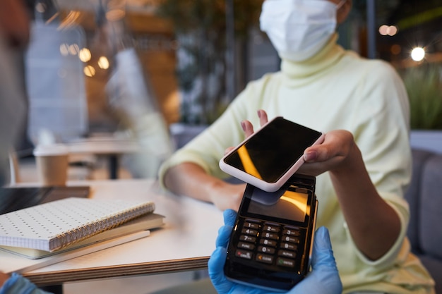 Cerca de irreconocible mujer con máscara pagando a través de smartphone en café con covid medidas de seguridad, espacio de copia