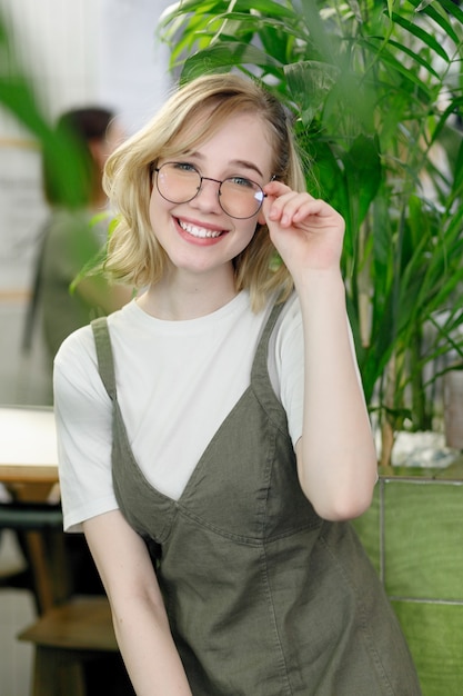 Cerca de joven alegre vestida felizmente mirando a puerta cerrada con grandes hojas verdes sobre fondo en un acogedor café verde. Niña, en, anteojos