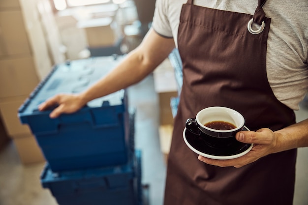 Cerca de joven en delantal sosteniendo una taza de bebida caliente y colocando la mano en la caja de plástico