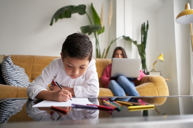 Foto cerca de la joven madre trabaja de forma remota en su computadora portátil mientras su hijo dibuja en un concepto de cuaderno de r ...