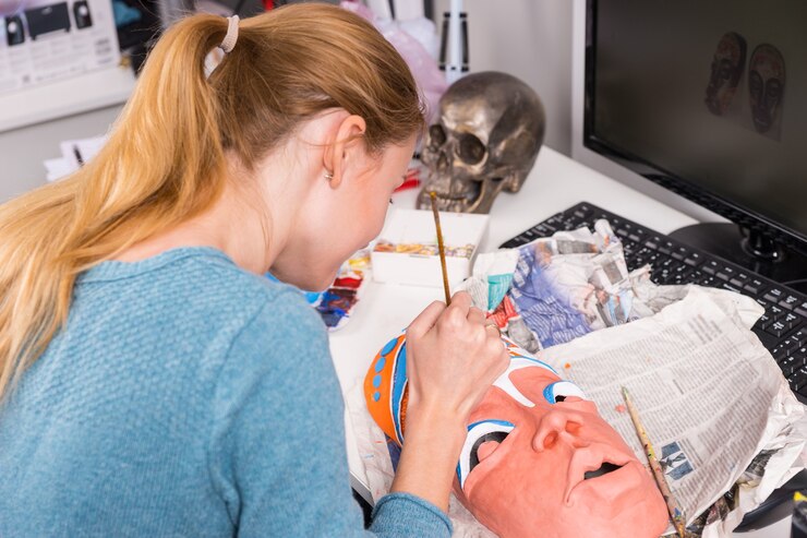 Foto cerca de la joven mujer artística rubia pintando detalles coloridos en la máscara tribal de arcilla mientras está sentado en el escritorio de la computadora