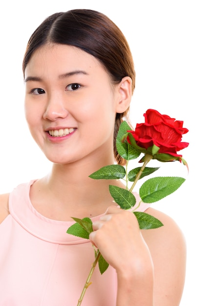 Cerca de la joven mujer asiática feliz sonriendo y sosteniendo una rosa roja cerca de la cara