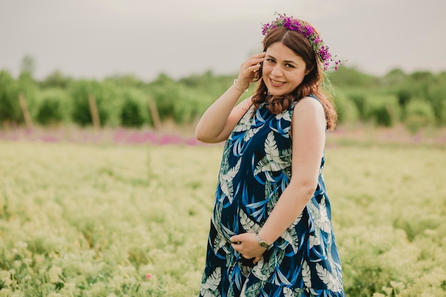 Cerca de la joven mujer embarazada sonriente con flores silvestres en la cabeza disfrutando de la maternidad de pie en el campo y mirando a la cámara