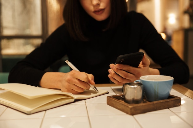 Foto cerca de una joven mujer morena sentada en la mesa de café en el interior, escribiendo en un cuaderno, sosteniendo el teléfono móvil